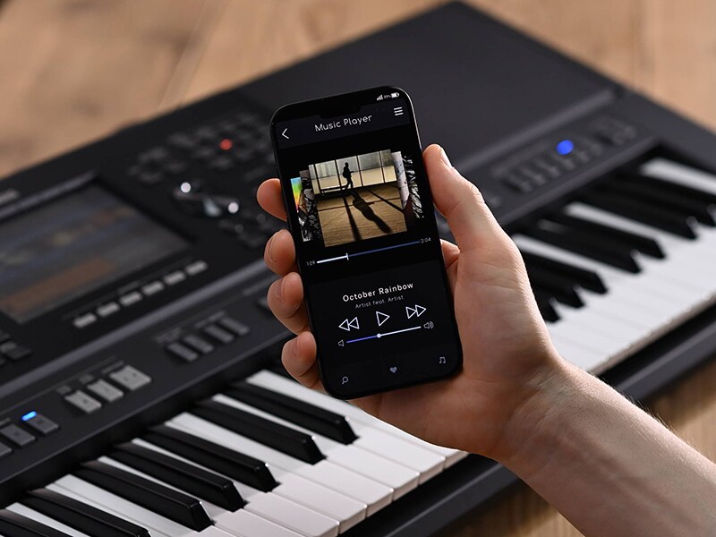 Yamaha Arranger Workstation PSR-SX920 en een hand die een smartphone vasthoudt met het scherm van een muziekspeler
