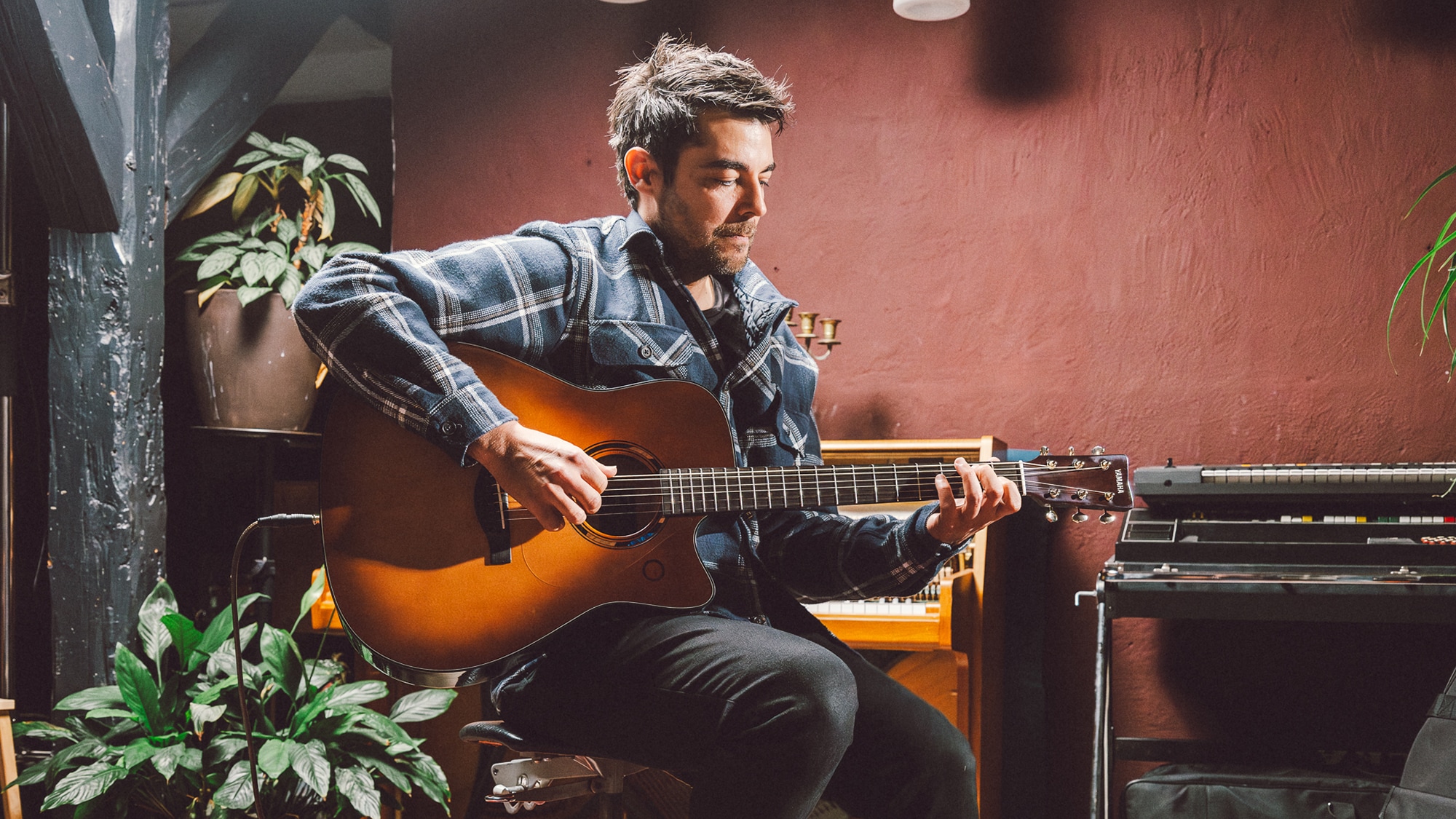 Man sitting by a piano and playing the TAG3 C acoustic guitar.
